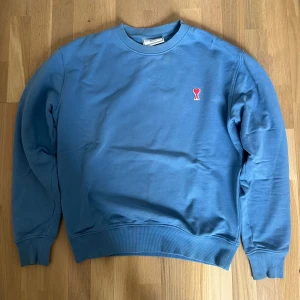 Ami Paris tröja - Säljer en blå sweatshirt från Ami Paris med klassisk rund hals och ribbade muddar. Tröjan har Ami:s ikoniska röda hjärta broderat på bröstet. Perfekt för dig som gillar stilren design och vill ha något bekvämt och snyggt.