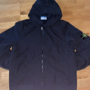 Svart vindjacka från Stone Island - Svart vindjacka från Stone Island med huva och dragkedja framtill. Jackan har den klassiska Stone Island-loggan på ärmen och två sidofickor med dragkedja. Perfekt för dig som gillar streetwear och vill ha en snygg jacka med coola detaljer.