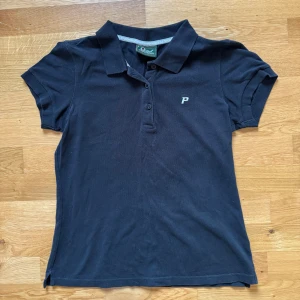 Svart pikétröja Peak Performance S - Klassisk svart pikétröja från Peak Performance i storlek S. Tröjan har korta ärmar, krage och knäppning framtill med tre knappar. Liten broderad P-logga på bröstet. Perfekt för en sportig och stilren look.