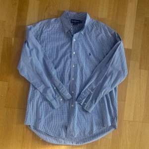 Rutig skjorta från Ralph Lauren - Snygg rutig skjorta från Ralph Lauren i blått och vitt. Klassisk modell med lång ärm, knappar framtill och broderad logga på bröstet. Tillverkad i 100% bomull för en skön känsla och stilren look.