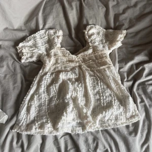 Vit blus med volangärmar - Säljer en söt vit blus med korta volangärmar och smockad struktur. Blusen har en v-ringad hals och är tillverkad i ett lätt och luftigt bomullsmaterial. Perfekt för varma dagar och ger en romantisk vibe till din outfit.