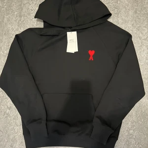 Svart hoodie från Ami med rött hjärta - Svart hoodie från Ami med klassisk huva och stor magficka. På bröstet finns Ami:s ikoniska broderade röda hjärta med A. Tröjan har ribbade muddar och är tillverkad i mjuk bomull, perfekt för chill dagar. Snygg och enkel design som funkar till allt.