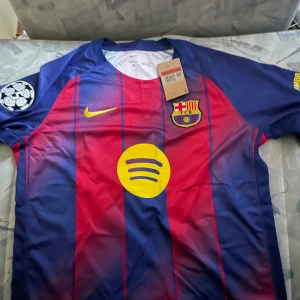 FC Barcelona Lamine Yamal matchtröja - Säljer en officiell FC Barcelona fotbollströja från Nike med Lamine Yamal och nummer 10 på ryggen. Tröjan har klassiska blå och röda vertikala ränder, gula detaljer och klubbmärke på bröstet. Materialet är lätt polyester, perfekt för match eller träning.