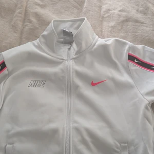Vit Nike track jacket med rosa detaljer - Nike track jacket i vitt med dragkedja framtill och höga krage. Snygga rosa och svarta detaljer längs ärmarna med Nike-loggor. Jackan har två fickor och är tillverkad i ett mjukt, sportigt material. Perfekt för en sportig och trendig look.