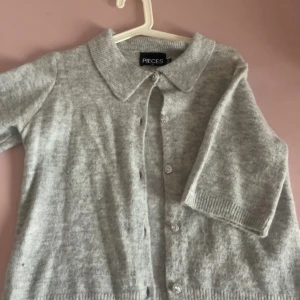 Grå kofta från Pieces med glittriga knappar - Superfin ljusgrå kofta från Pieces i stickad kvalitet. Klassisk krage och långa ärmar, dekorerad med glittriga knappar som ger en extra touch. Perfekt att slänga över en topp eller klänning för en snygg layering.