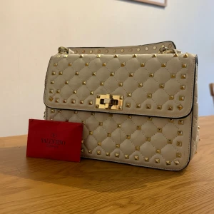 INTRESSEKOLL Valentino Garavani - Säljer en vit Valentino Garavani rockstud med guld nitar. Kan inte garantera äktheten då den är köpt här på Plick och fick inte med något kvitto, men fick med ett kort (se bild). Fint skick men använd🩷 Säljer då jag inte använder den längre🩷