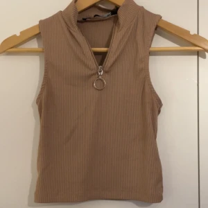 Beige ribbad ärmlös topp med dragkedja - Säljer en beige ribbad ärmlös topp med hög krage och snygg dragkedja framtill med rund metallring. Perfekt för en trendig och avslappnad look. Passar till både jeans och kjol.