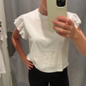 Vit blus med volangärmar - Säljer en söt vit blus med korta volangärmar. Toppen har en rund halsringning och är tillverkad i mjuk bomull. Perfekt för dig som gillar en clean och fräsch look med lite extra detaljer på axlarna.