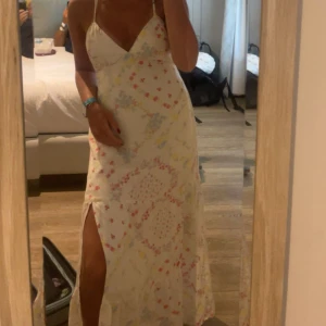 Vit långklänning med blommigt mönster - Supersöt vit långklänning med tunna axelband och v-ringning. Klänningen har ett blommigt mönster i ljusblått, gult och rött samt en slits på sidan för extra vibe. Perfekt för varma dagar och har en flowy, loose passform.