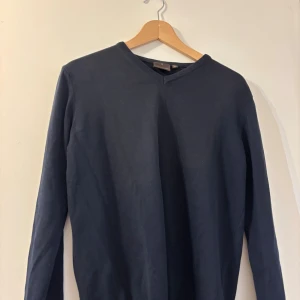 Mörkblå v-ringad tröja Oscar Jacobson - Snygg mörkblå v-ringad tröja från Oscar Jacobson, tillverkad i Italien. Tröjan har lång ärm och ribbade detaljer vid halsringning, ärmslut och nederkant. Perfekt för dig som gillar stilrena och klassiska plagg med en modern touch.