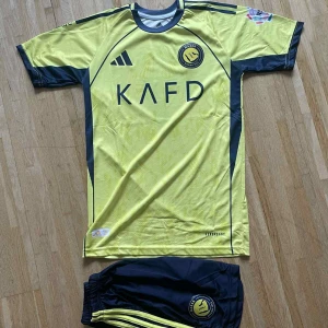 Al Nassr Ronaldo 7 matchtröja Adidas - Gul och mörkblå Al Nassr fotbollströja med Ronaldo 7 på ryggen. Tröjan har korta ärmar, klubbmärke och sponsortryck framtill samt Adidas-logga. Tillverkad i lätt och ventilerande material, perfekt för fotbollsträning eller match.