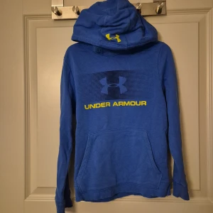 Blå Under Armour hoodie loose fit - Blå hoodie från Under Armour med gul logga och text framtill. Tröjan har stor huva, magficka och ribbade muddar. Loose fit för skön och avslappnad stil. Perfekt för träning eller chill. Materialet är mjuk bomull och polyester. Skulle säga storlek 140/146