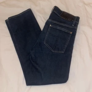 Tiger of Sweden Iggy jeans blå 30/32 - Snygga blå jeans från Tiger of Sweden, modell Iggy. Klassisk femficksdesign med silverfärgade nitar och mörkblå tvätt. Raka ben och normal passform, tillverkade i slitstarkt jeanstyg. Perfekta för dig som gillar stilrena och tidlösa jeans.