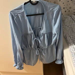 Ljusblå blus med snörning från AWARE - Supersöt ljusblå blus från AWARE med volanger och två snörningar framtill. Blusen har långärmade ärmar med rynkade muddar och en djup v-ringning. Materialet är mjukt och har en lätt glansig finish som ger en chill vibe. Perfekt för att lyfta din look!