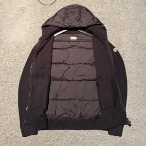 Moncler cardigan  - Svart pufferjacka från Moncler med stickade ärmar och sidor, quiltad front och rygg samt stor huva. Jackan har dragkedja och ribbade muddar för extra komfort. Perfekt för kyliga dagar när du vill vara både varm och snygg. Passar även S