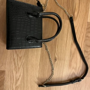 Svart handväska med guldkedja - Snygg svart handväska i med guldfärgad kedja och dragkedja upptill. Väskan har både handtag och axelrem. Har en liten defekt vilket är handtaget är böjt men annars funkar den som vanligt!