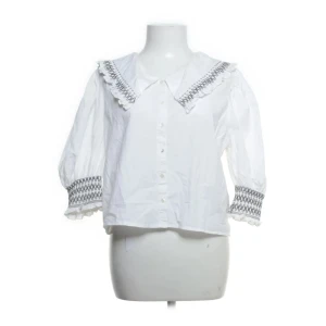 Vit blus med brodyr från Zara - Supersöt vit blus från Zara med bred krage och broderade detaljer i svart på både krage och ärmslut. Blusen har trekvartsärmar med volangkant och knäppning framtill. Perfekt för dig som gillar romantisk och trendig stil.