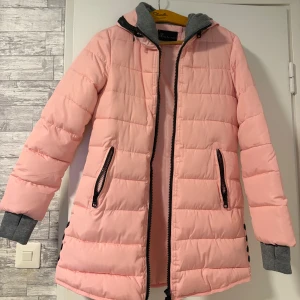 Ljusrosa pufferjacka från Xin bo xui - Säljer en ljusrosa pufferjacka från Xin bo xui med svarta dragkedjor och gråa muddar. Jackan har huva, två fickor med dragkedja och snygga knappar på sidan. 