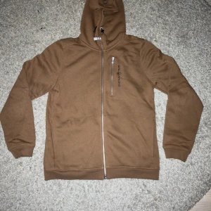 Brun hoodie från Sail Racing med dragkedja - Säljer en brun hoodie från Sail Racing med hel dragkedja och en extra dragkedja på bröstet. Tröjan har huva, ribbade muddar och är tillverkad i mjuk bomull. Perfekt för dig som gillar stilren design och vill ha något bekvämt att slänga på dig.