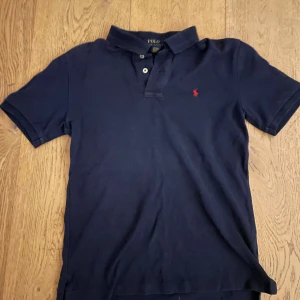 Marinblå pikétröja från Polo Ralph Lauren - Snygg marinblå pikétröja från Polo Ralph Lauren med klassisk krage och två knappar framtill. Tröjan har korta ärmar och den ikoniska röda logon broderad på bröstet. Tillverkad i 100% bomull för skön och luftig känsla. 