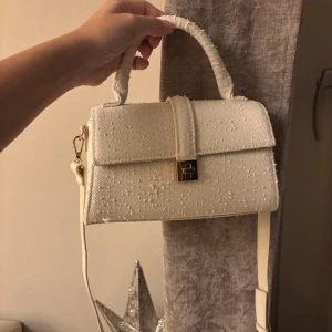 Vit handväska från H&M - Stilren vit handväska med snygg struktur i tyget och guldfärgat spänne framtill. Väskan har både handtag och avtagbar axelrem, vilket gör den enkel att bära på olika sätt. 