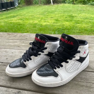 Nike Jordan 1 Access st 37,5 - Säljer ett par Nike Jordan 1 sneakers i svart och vitt med röda Jumpman-detaljer på plösen. Skorna har klassisk snörning, perforerade paneler och rund tå. Tillverkade i skinn med svart platt sula och cool high-top siluett. Perfekt för dig som gillar streetstyle.