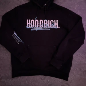 Svart hoodie från Hoodrich med tryck - Svart hoodie från Hoodrich i storlek M med stor vit och blå logga på bröstet och texten 'From nothing to something' på ryggen och ärmen. Tröjan har huva med snörning och en klassisk magficka. Perfekt streetwear med coolt grafiskt tryck.