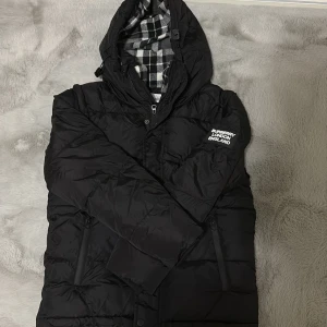 Svart dunjacka från Burberry - Säljer en svart dunjacka från Burberry med huva och dragkedja. Jackan har ett rutigt foder i svart och vitt, samt Burberry London England-tryck på bröstet. Fylld med dun och fjäder, yttertyg i polyester. Perfekt för kalla dagar.