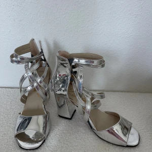 Silvriga sandaler med klack och remmar - Snygga sandaler i metallic silver med bred klack och flera smala remmar som korsar över foten och runt ankeln. Skorna har öppen tå och en glansig finish som verkligen sticker ut. Perfekta för dig som vill ha något extra till outfiten.