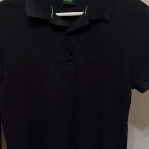 Svart pikétröja från BOSS - Säljer en svart pikétröja från BOSS med diskret broderad logga på bröstet. Tröjan har klassisk krage, knäppning framtill och snygga gröna detaljer på insidan av kragen. Perfekt för en clean och stilren look.