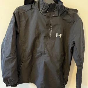 Svart vindjacka från Under Armour - Svart vindjacka från Under Armour med huva och diskret logga på bröstet. Jackan har dragkedja framtill, elastiska muddar och är perfekt för blåsiga dagar. Snygg och sportig look med stilrena detaljer. Storlek M men passar S,Aldrig använd