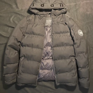 Svart pufferjacka från Supply & Demand - Säljer en svart pufferjacka från Supply & Demand med huva och dragkedja. Jackan har en patch på ärmen, två fickor med dragkedja och breda, vadderade paneler. Perfekt för kalla dagar och riktigt snygg streetstil.