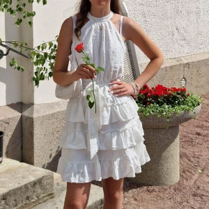 Vit volangklänning med knyt i midjan - Superfin vit kortklänning med volanger och knyt i midjan. Klänningen är ärmlös och har ett lätt prickigt mönster i tyget. Perfekt för sommarens alla tillfällen och ger en romantisk vibe.
