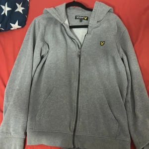Grå hoodie från Lyle & Scott Junior - Säljer en grå hoodie från Lyle & Scott Junior med dragkedja och huva. Tröjan har ett broderat gult logomärke på bröstet, två fickor framtill och är långärmad. Perfekt för chill dagar och enkel att matcha med jeans eller joggers. Storlek 14-15 år 