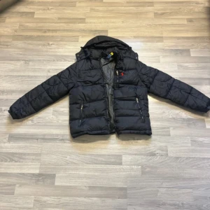Svart pufferjacka från Polo Ralph Lauren - Svart pufferjacka från Polo Ralph Lauren med rött broderat logomärke på bröstet. Jackan har huva, två framfickor med dragkedja och quiltad design. Materialet är polyester och jackan stängs med dragkedja framtill. Perfekt för kalla dagar. Helt ny! 