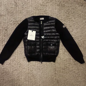 Svart Cardigan Moncler  - Storlek S i prencip helt ny. Har kvitto! Säljer en cardigan pufferjacka från Moncler med quiltad front och två fickor med knappar. Jackan har långärmade stickade ärmar och dragkedja framtill. Klassisk Moncler-logga på ärmen och ribbad krage. Perfekt för dig som vill ha en stilren och trendig look.
