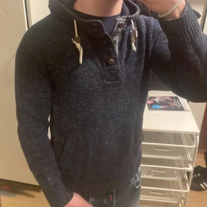 Blå stickad hoodie - Fin blå stickad hoodie med knappar vid kragen. Passar otroligt bra nu till kallare väder. Priset kan diskuteras. Modellen är 180cm, 73kg.