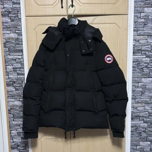 Svart dunjacka från Canada Goose - Säljer en svart dunjacka från Canada Goose med klassisk patch på ärmen. Jackan har huva, dragkedja och knappar framtill samt dragsko i nederkant. Perfekt för kalla vinterdagar och riktigt snygg streetstil.