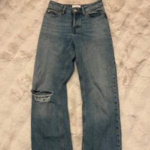 Blå raka jeans med slitning på knät - Säljer ett par klassiska blå jeans med rak passform och medel hög midja. Jeansen har en snygg slitning på ena knät. Perfekt för dig som gillar en avslappnad och trendig look.