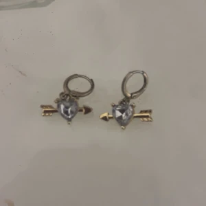 Guldiga örhängen med hjärta och pil  - Supersöta örhängen i guldton med glittrande hjärtformad sten och pil-detalj. Perfekta för dig som vill ha något unikt och lekfullt i öronen. Hängande modell som verkligen sticker ut. 