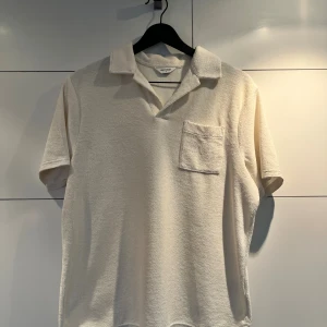 Beige pikétröja i frotté från East West - Säljer en beige pikétröja från East West i mjuk frotté. Tröjan har klassisk krage, kort ärm och en bröstficka. Perfekt för chill vår- och sommardagar när du vill ha en avslappnad men ändå clean look.