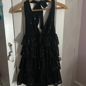Svart glittrig volangklänning med rosett - Supersnygg svart kortklänning med volanger och paljetter. Djup v-ringning fram och öppen rygg som knyts med en bred rosett i nacken. Perfekt för dig som vill sticka ut och glänsa! Använd 1 gång på nyår förra året ‼️