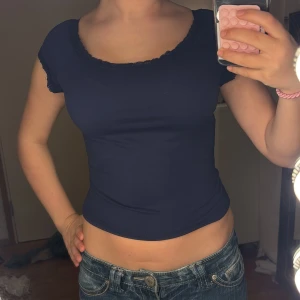 Mörkblå croppad topp med spets - Snygg mörkblå croppad topp med korta ärmar och spetsdetaljer vid halsringning och ärmslut. Toppen har en tight passform och är tillverkad i mjuk bomull som sitter skönt mot kroppen. Perfekt för dig som gillar stilrena och feminina plagg.