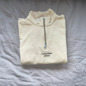 Beige half zip sweatshirt Calvin Klein - Snygg beige sweatshirt från Calvin Klein Jeans med half zip och CK-detalj på dragkedjan. Tröjan har en stor magficka, ribbade muddar och broderad logga på bröstet. Perfekt för chill dagar och enkel att matcha med jeans eller joggers.