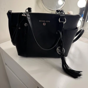 Svart handväska från Michael Kors - äkta!!!!!! ordinarie:3000kr                                                        Snygg svart handväska från Michael Kors med silverfärgade detaljer och logotyp framtill. Väskan har både handtag och axelrem, samt en dekorativ tofs med MK-hänge. Stilren och rymlig modell i skinn med dragkedja upptill och praktiska fack på sidorna.