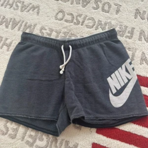 Svarta shorts från Nike med snörning - Svarta mjuka shorts från Nike med stor vit logga på ena benet. De har en bekväm midja med vit snörning och råa kanter vid bensluten. Perfekta för chill eller träning, klassisk stil och loose passform.
