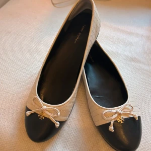 Tory Burch ballerinaskor beige & svart - Snygga ballerinaskor från Tory Burch i beige med svarta tår. Skorna har rund tå, dekorativ rosett framtill och guldfärgad logga. Klassisk och stilren design som passar till allt. Använt skorna 1 gång. 