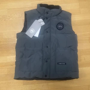 Grå dunväst från Canada Goose - Snygg grå dunväst från Canada Goose med hög krage och svarta knappar framtill. Västen har ett broderat svart logomärke på bröstet och en ficka med Canada Goose-etikett. Perfekt för kalla dagar och har en clean, modern look.