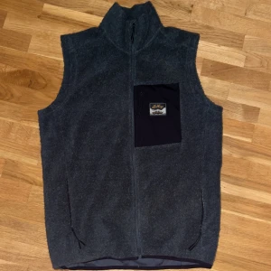 Olivgrön ullväst från Lundhags - Olivgrön väst från Lundhags i mjuk ullmix med hög krage och hel dragkedja framtill. Praktisk bröstficka med dragkedja och Lundhags-logga, samt två sidofickor med dragkedja. Riktigt varm skulle jag vilja säga. Perfekt för lager-på-lager och outdoor.