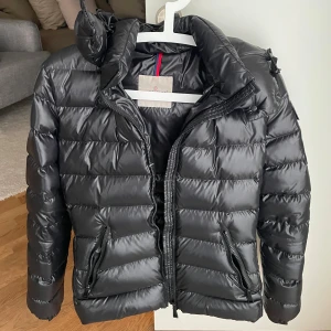 Svart Moncler pufferjacka - Svart pufferjacka från Moncler med glansig finish och huva. Jackan har quiltad design, dragkedjor på fickorna. Perfekt för kalla dagar. Passformen är ganska tajt då det är modellen Bady. Den är köpt 2021 och inte använd så många gånger så den är fortfarande i nyskick. Jag skickar gärna fler bilder och svarar på frågor om du är intresserad.